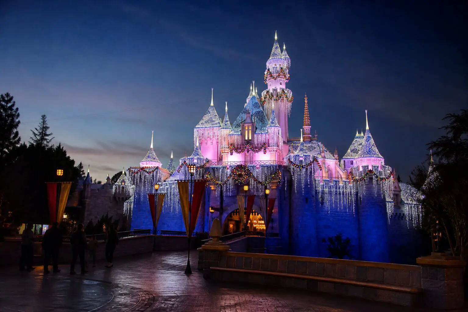 Sleeping Beauty Castle at Disneyland at dusk with winter icicle lights, wreaths, and a few guests on the castle bridge