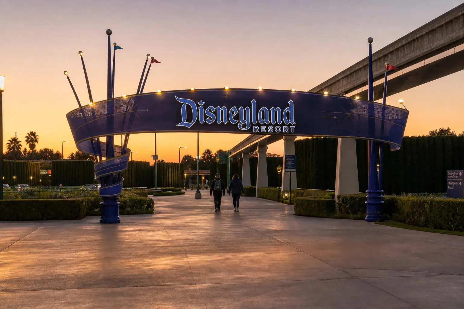 Disneyland Resort pedestrian entrance sign and walkway from the parking structures at dusk, with two guests walking under the arch and the monorail beam on the right